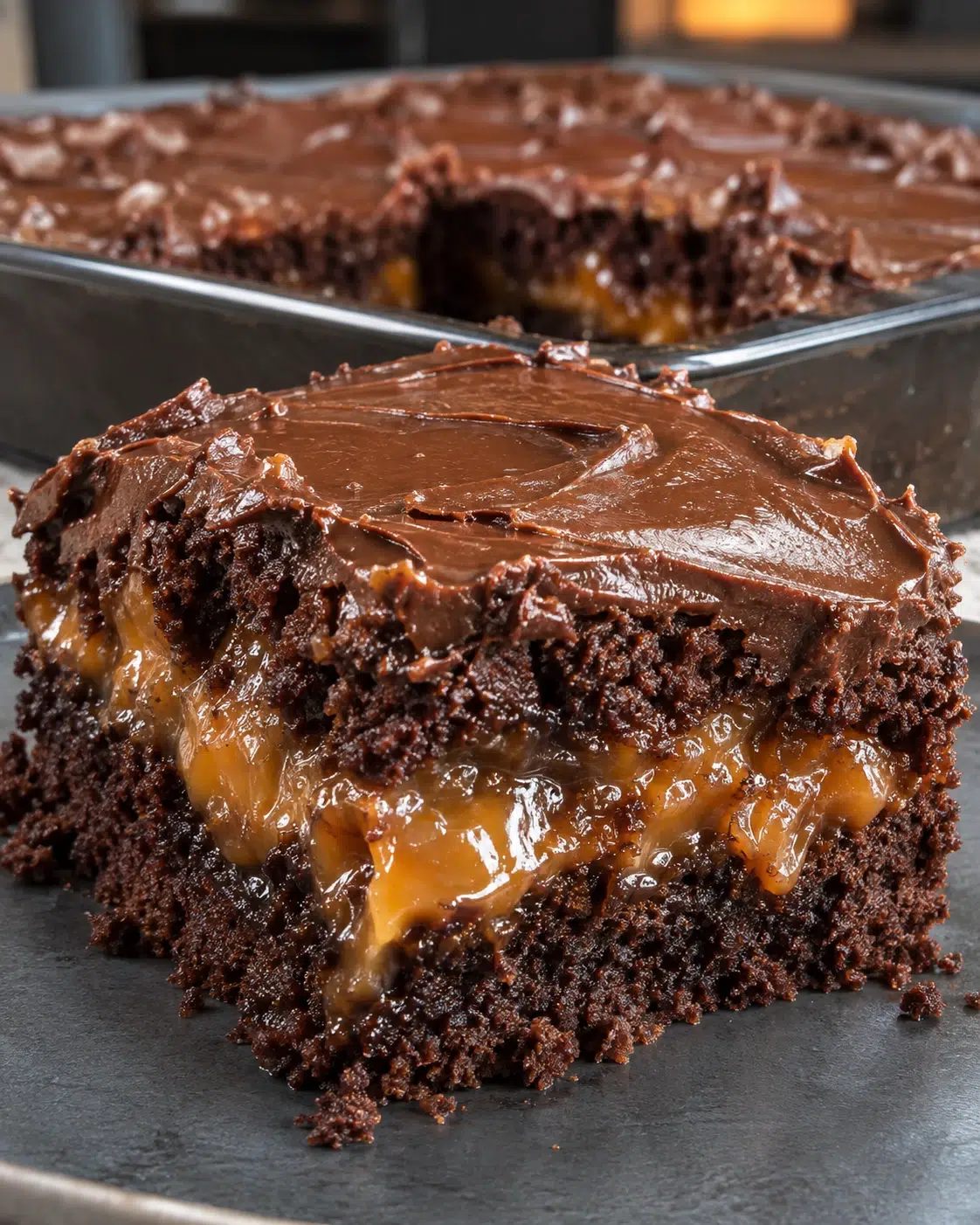 Sugar-free chocolate caramel cake bar with a glossy caramel middle layer and thick swirled chocolate frosting, in front of the pan of cake bars